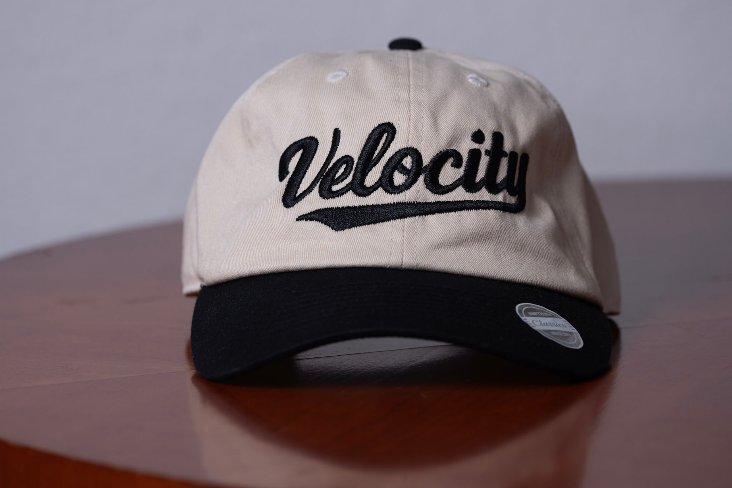 Beige and black baseball cap with 'Velocity' embroidered on the front and a sticker on the brim, placed on a wooden surface.