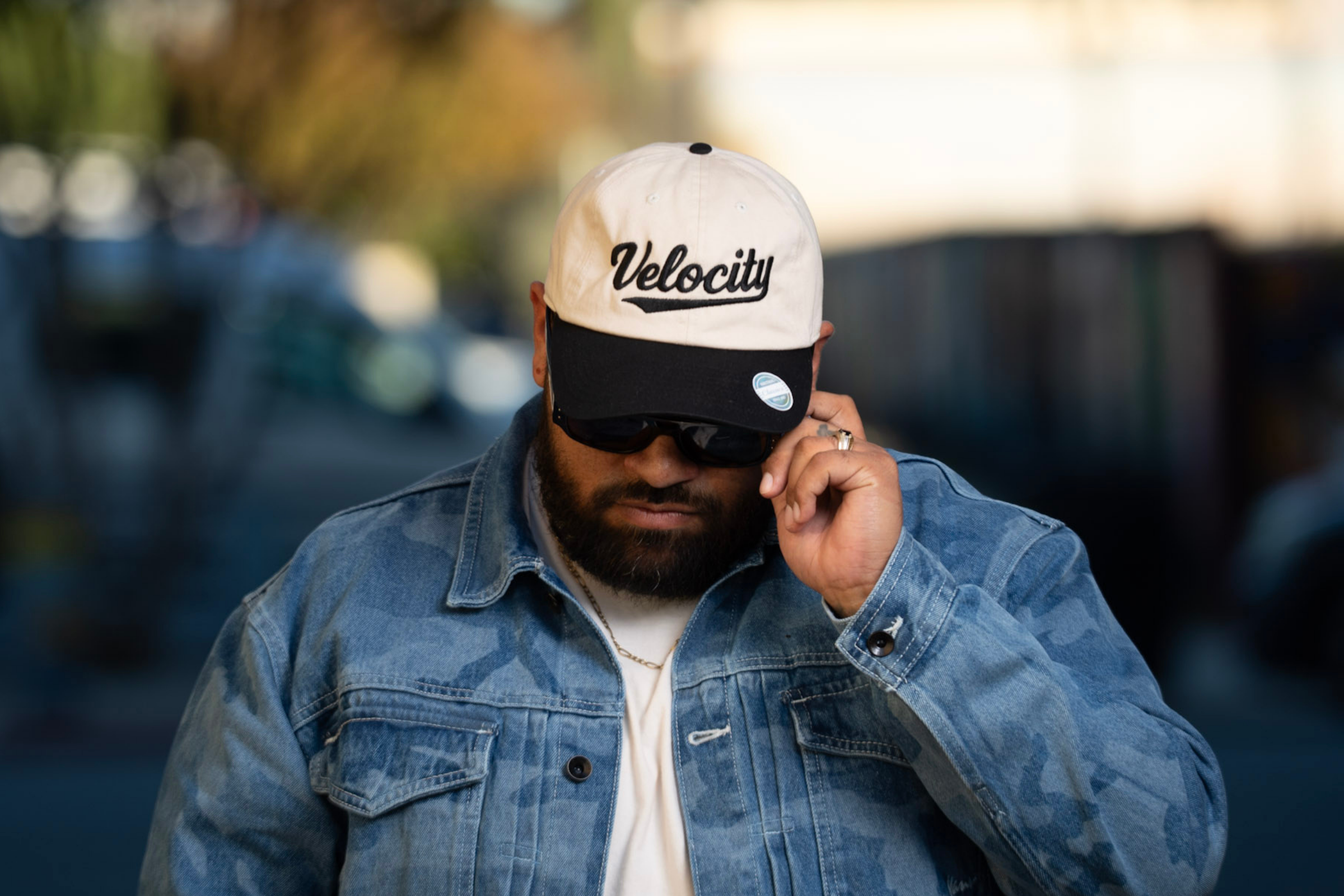 A man wearing a white and black cap with the word 'Velocity', dark sunglasses, a denim jacket, and a white shirt, adjusting his sunglasses outdoors.