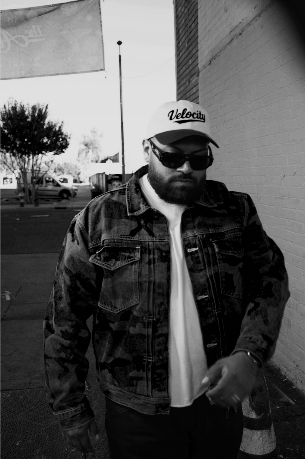 A man with a beard wearing sunglasses, a baseball cap with 'Velocity' written on it, a camouflage jacket, and a white shirt stands on a sidewalk near a brick wall.