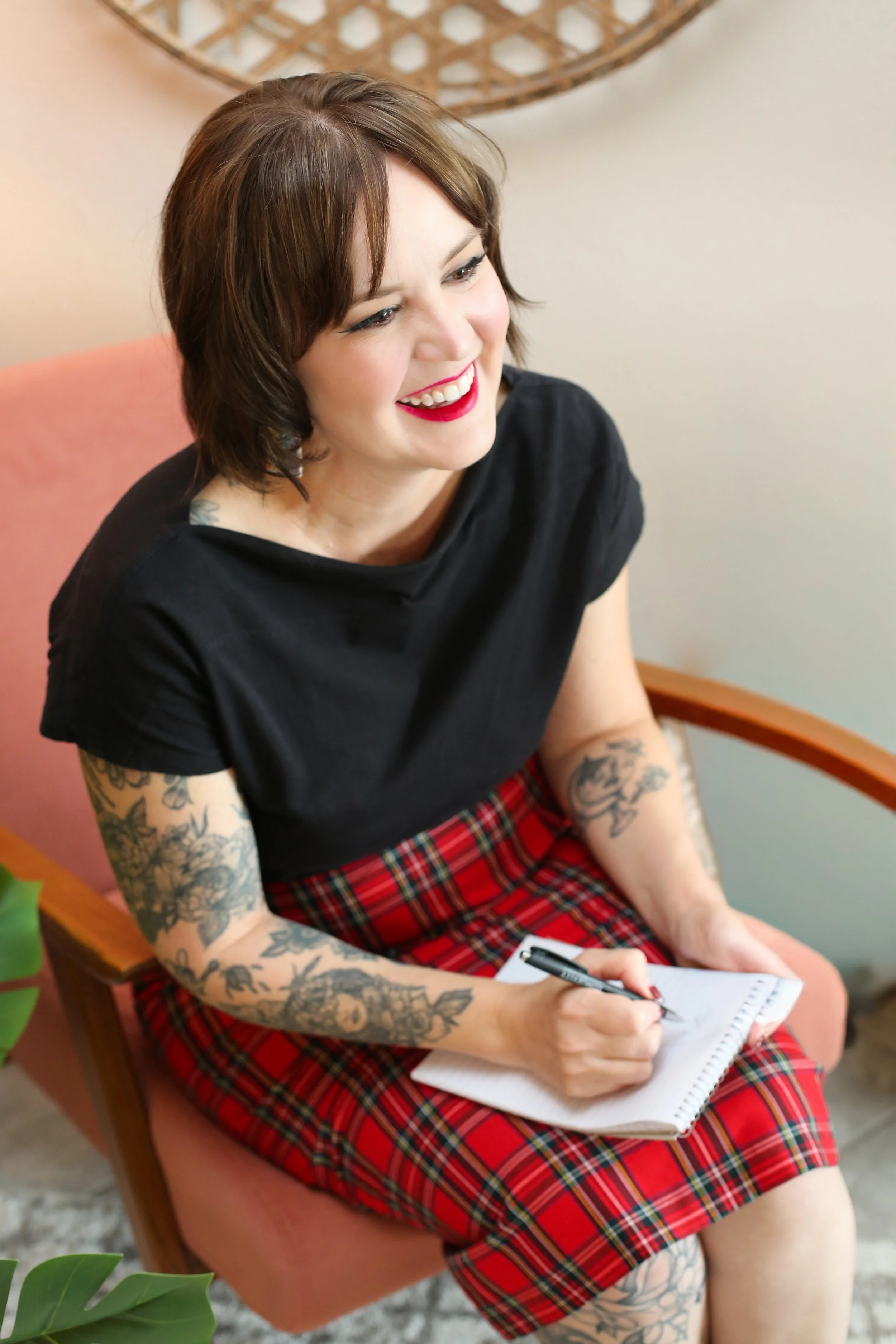 A woman with short brown hair, tattoos on her arms, wearing a black top and a red plaid skirt, sitting on a pink chair, smiling, holding a notepad and pen, in a cozy indoor setting.