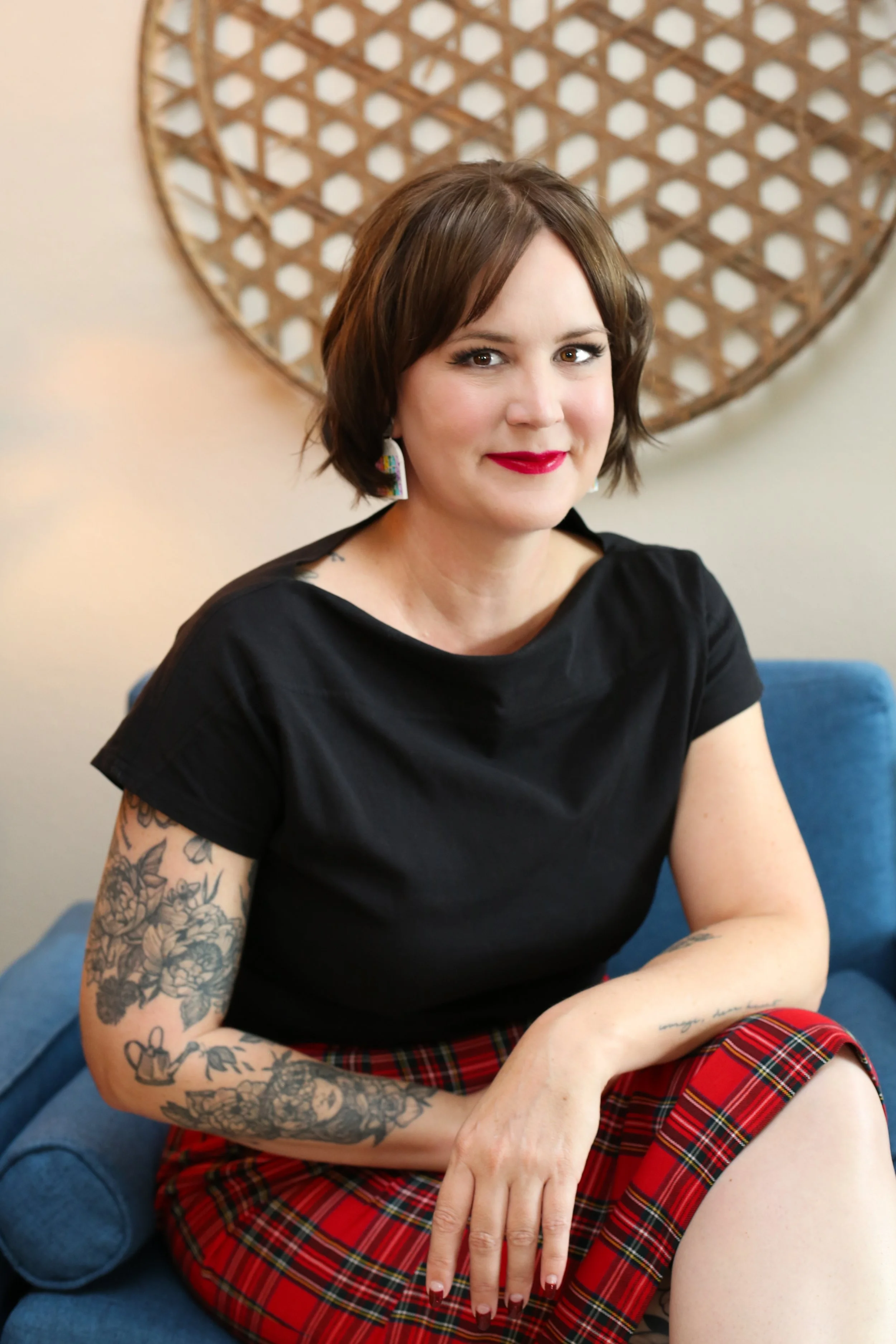 A woman with short brown hair, wearing a black top and a red plaid skirt, sitting on a blue couch. She has tattoos on her arms and is smiling at the camera, with a decorative wooden wall hanging behind her.