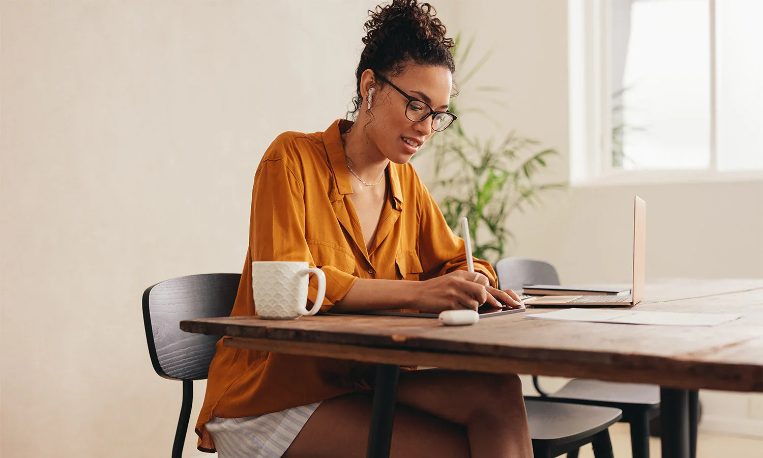 Woman with glasses and curly hair, wearing a mustard-colored shirt, sitting at a wooden table working with a pen, with a coffee mug, laptop, and notebook on the table, in a bright room with large windows and a potted plant in the background.