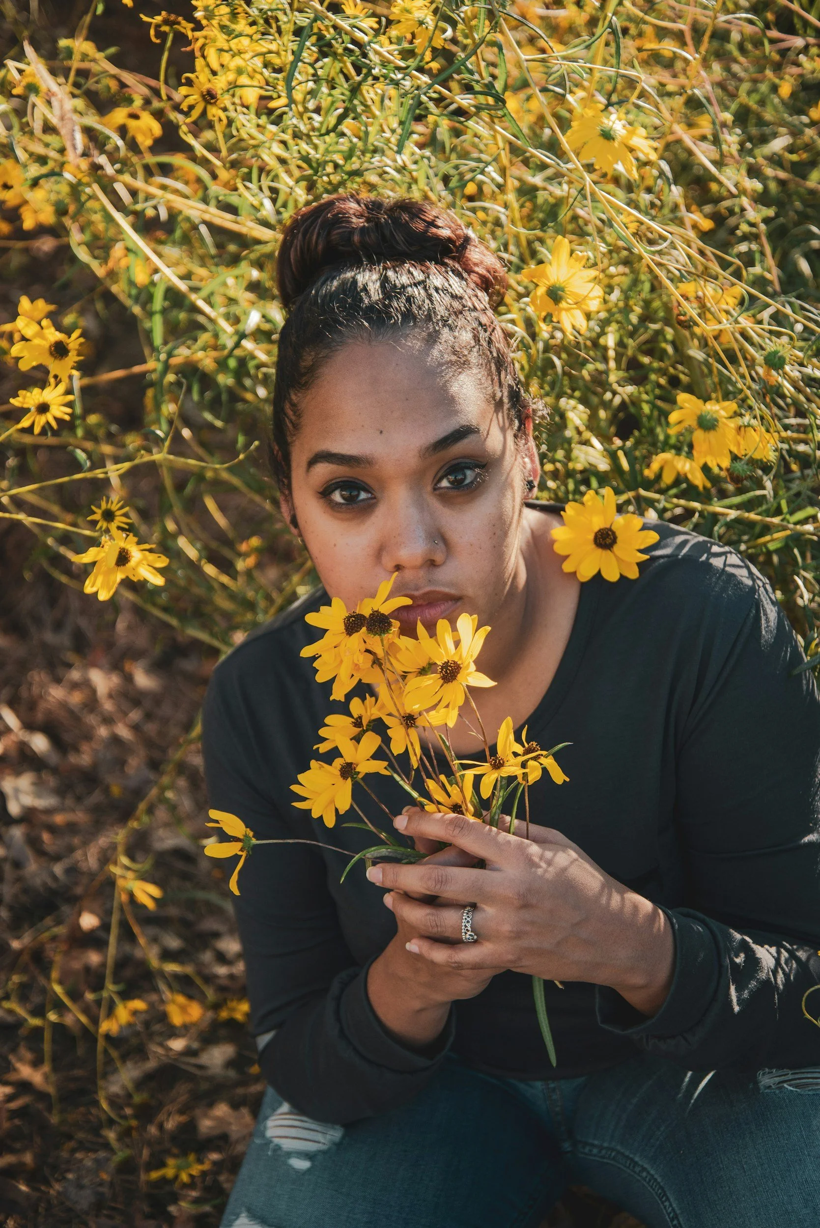 A woman with dark hair in a bun holding yellow flowers, sitting outdoors surrounded by more yellow flowers and greenery.
