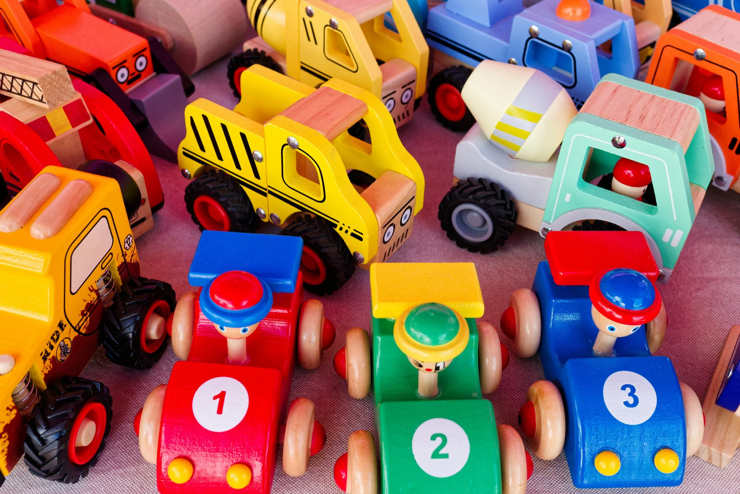 Colorful wooden toy vehicles, including race cars numbered 1, 2, and 3, and construction vehicles such as dump trucks and excavators, arranged on a surface.