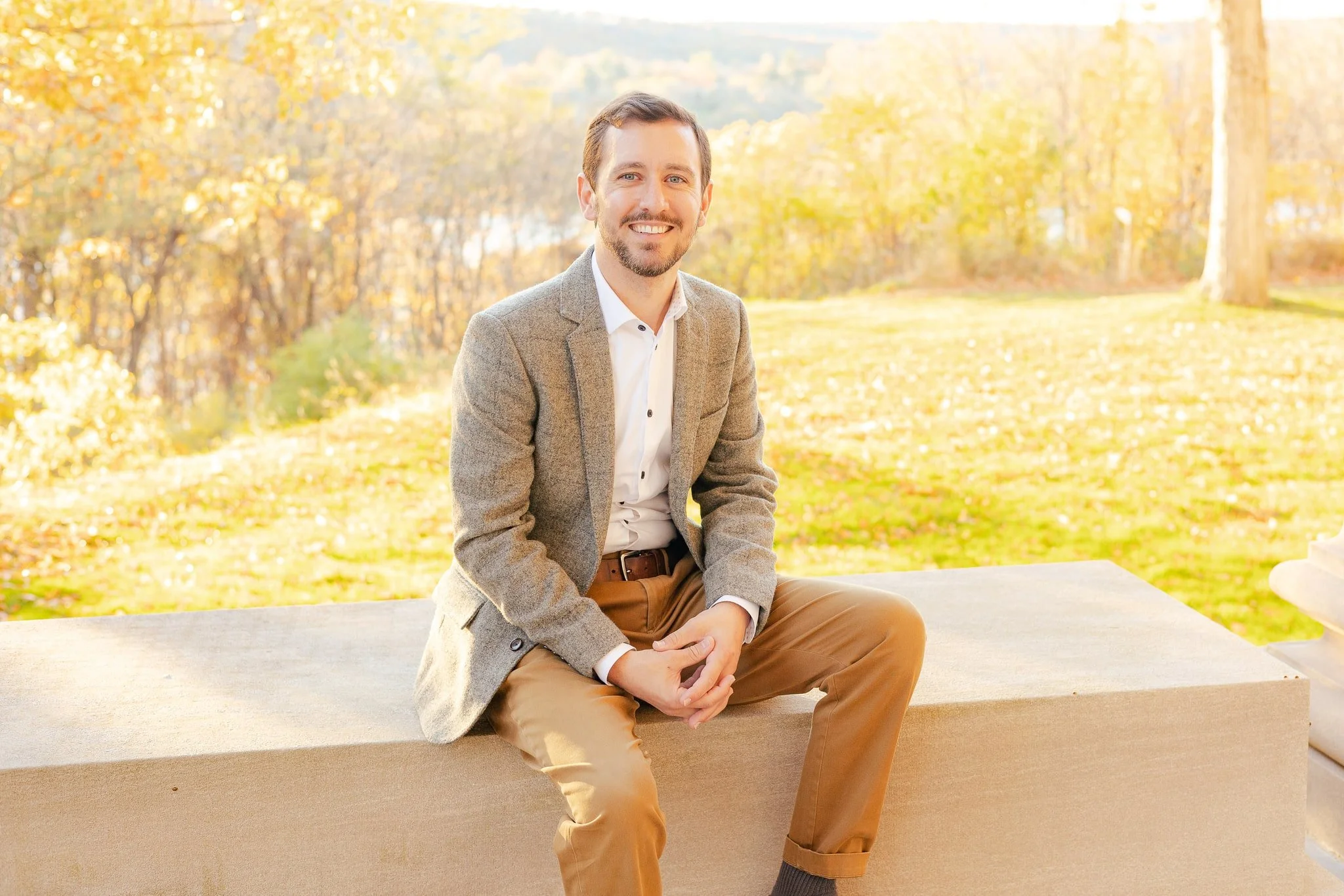 A smiling man in a beige blazer and tan pants sitting outdoors on a stone bench against an autumn background with trees and golden leaves.