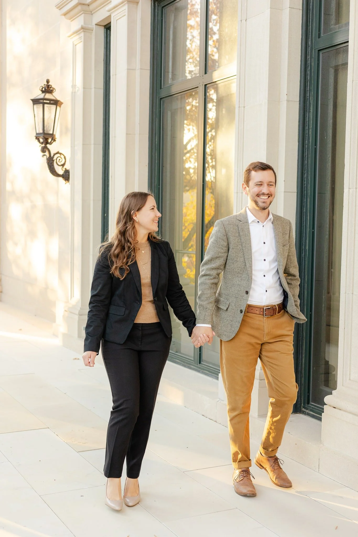 A smiling man and woman holding hands walking past a building with large windows and vintage exterior lighting.