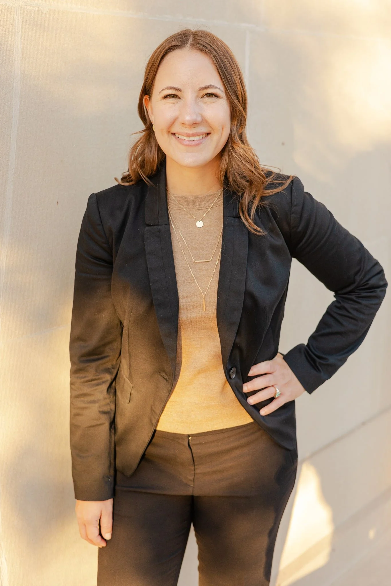 A woman with long brown hair, wearing a black blazer, beige top, and black pants, standing outdoors against a light-colored wall, smiling with one hand on her hip.