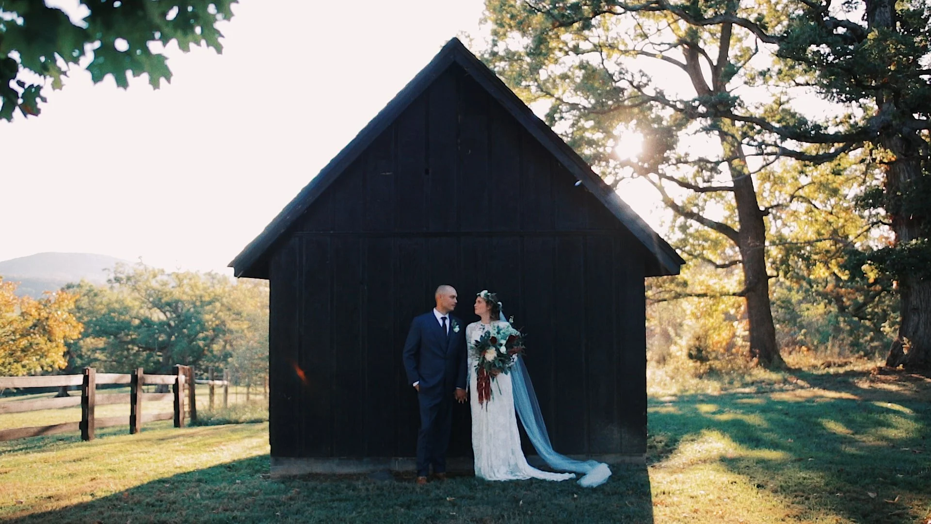 A bride and groom standing hand in hand outdoors at sunset, with a dark wooden barn in the background and trees around, during their wedding.