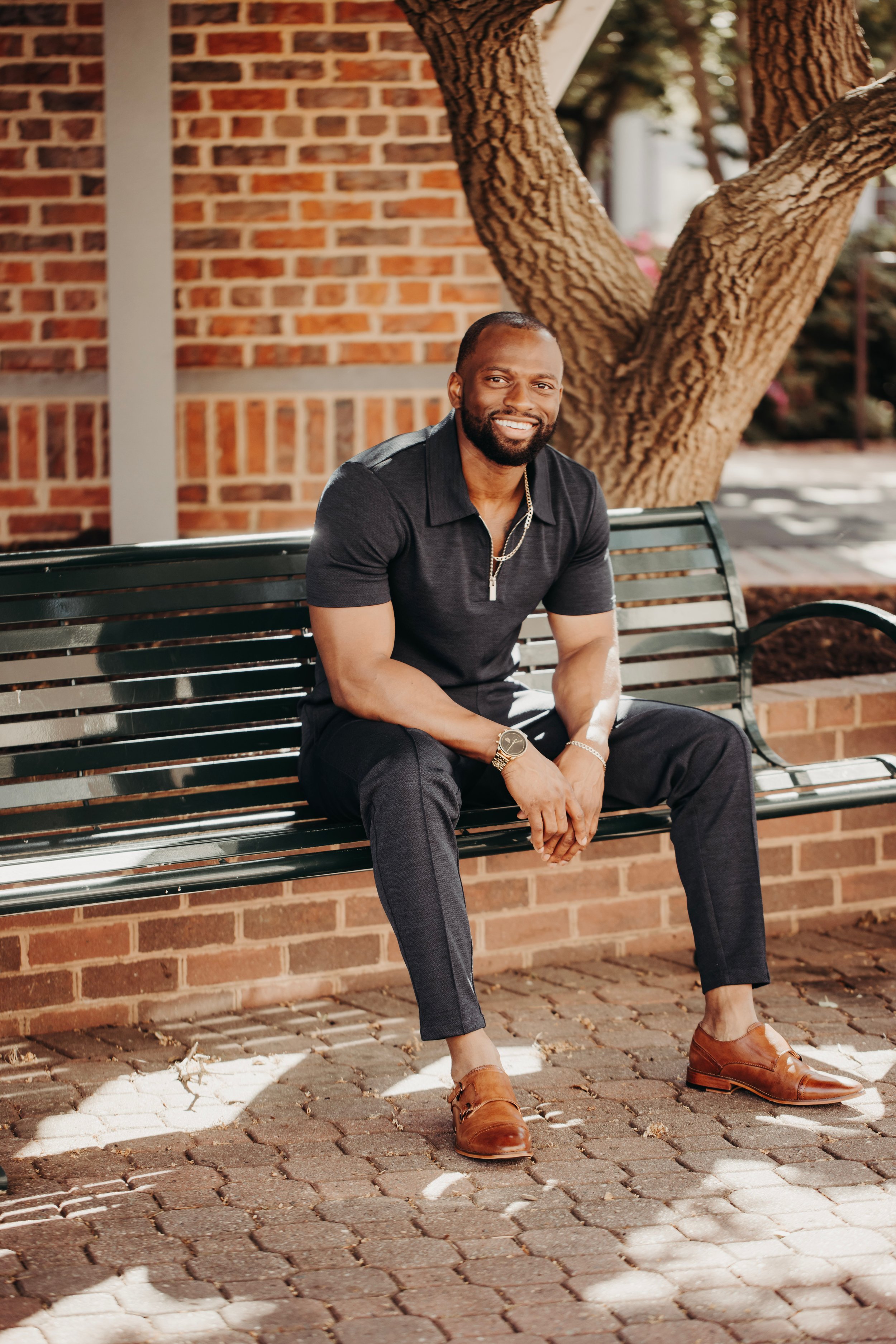 A smiling man with a beard and short hair, wearing a black polo shirt, dark dress pants, brown dress shoes, and accessories, sitting on a park bench under a tree with brick wall background.