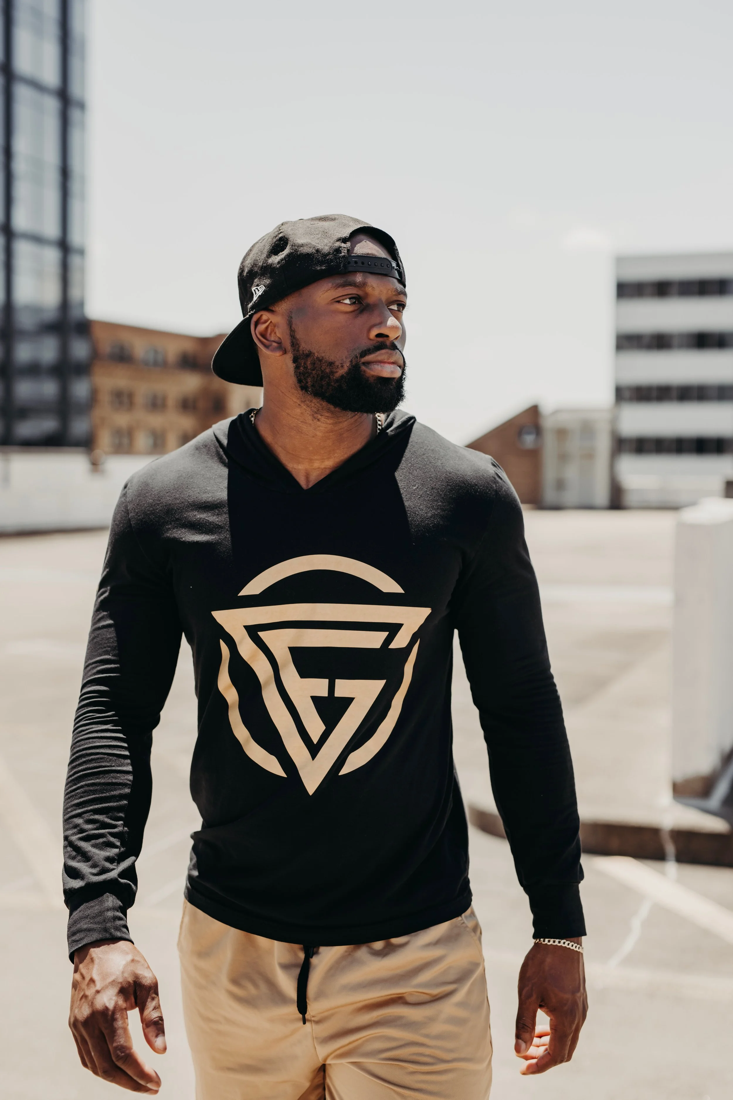 A young man with a beard and black cap backwards wearing a black long sleeve shirt with a geometric logo, beige shorts, standing outdoors on a rooftop with city buildings in the background.
