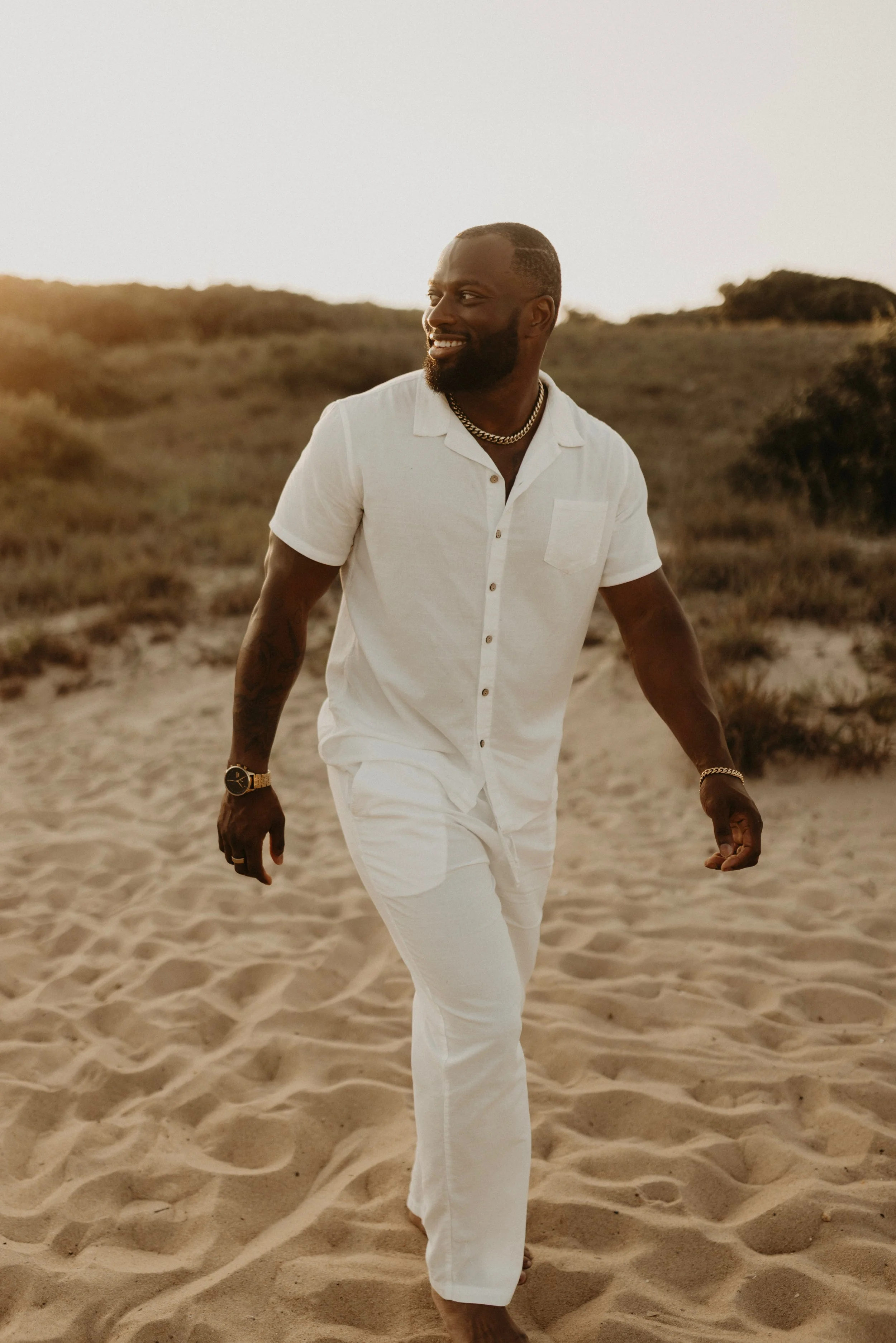 A man walking on sandy beach during sunset, dressed in white shirt and pants, smiling, wearing gold accessories.