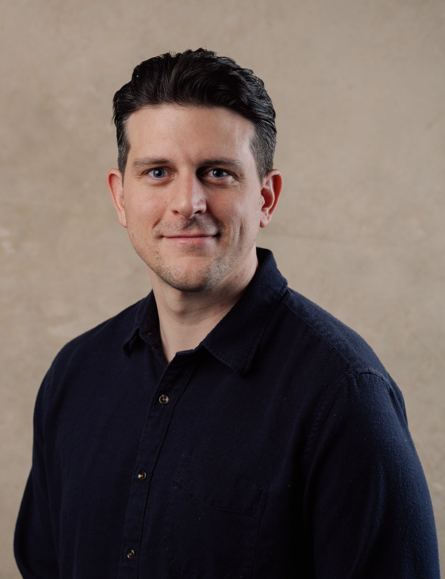 Headshot of a man with dark hair, light skin, wearing a black collared shirt, against a beige background.
