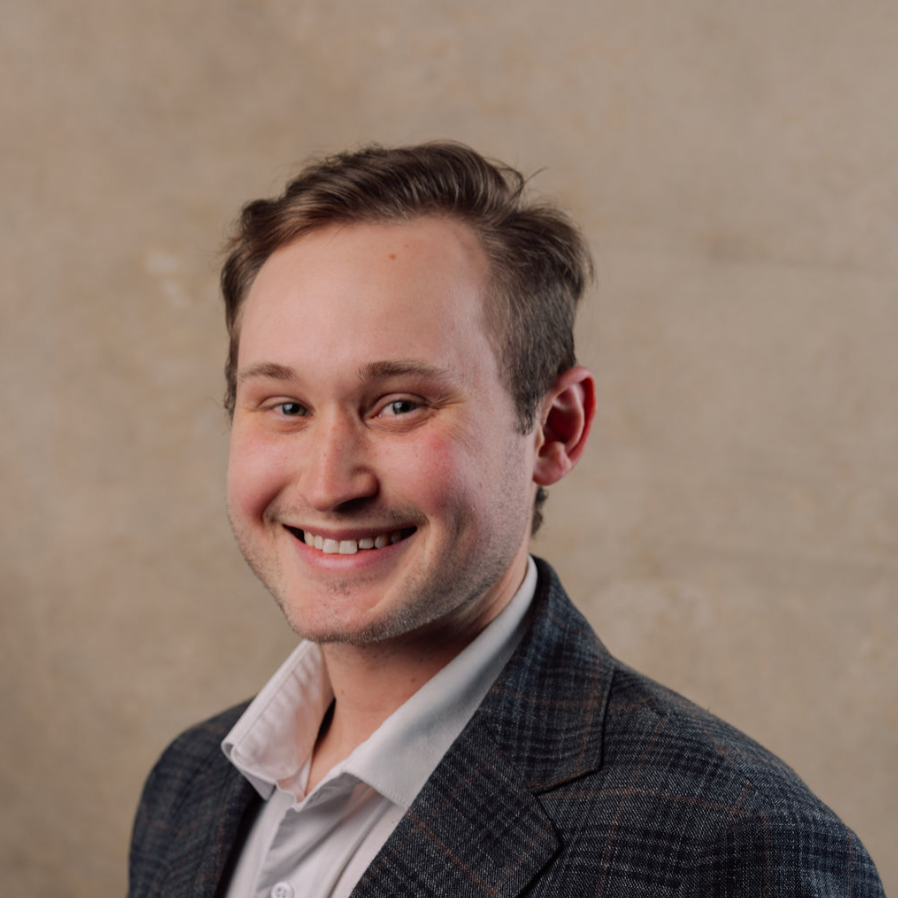 Portrait of a smiling young man with short brown hair, wearing a dark checkered blazer and white shirt, against a beige textured background.