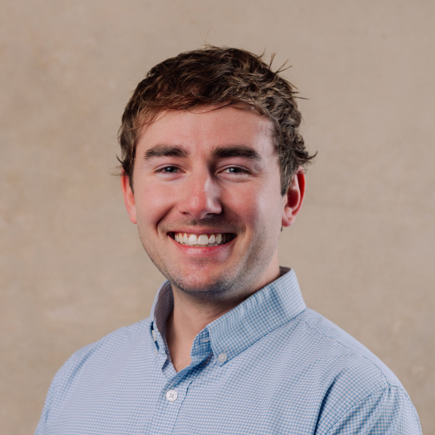 Young man with light brown hair, smiling, wearing a blue checkered button-up shirt, against a beige background.