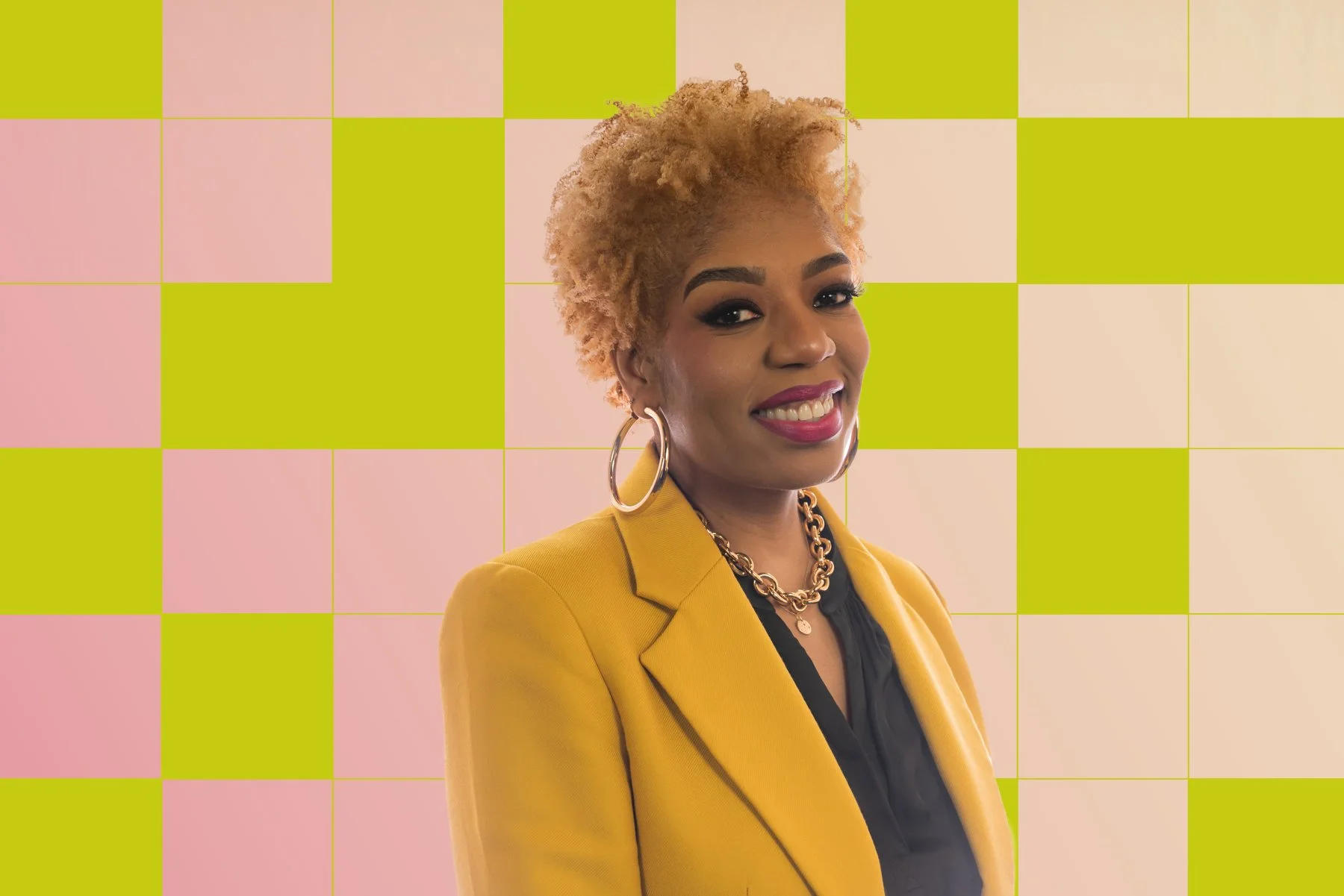 Portrait of a woman with short curly blonde hair, wearing a yellow blazer, gold hoop earrings, and a gold chain necklace, smiling against a background of pink and green checkered pattern.