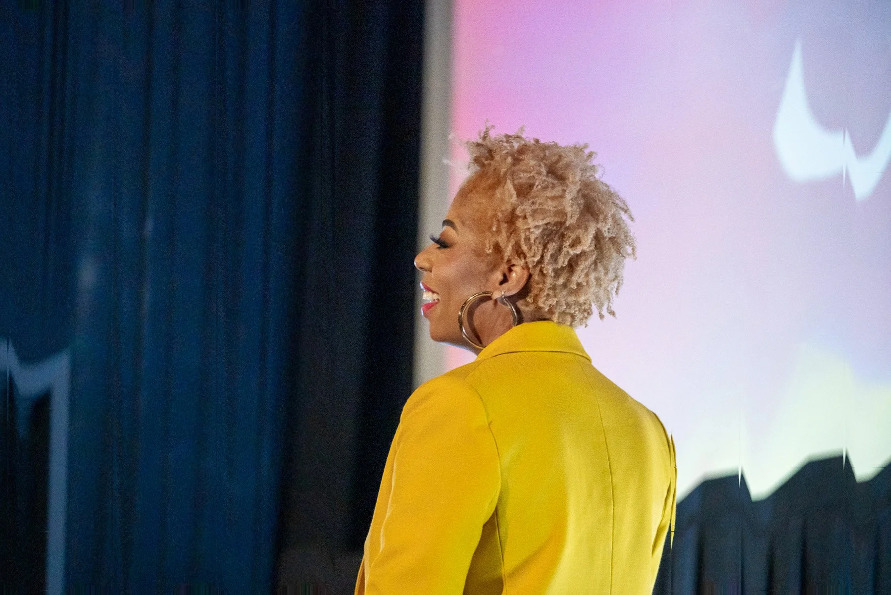 A woman with curly blonde hair, wearing large hoop earrings and a yellow blazer, is smiling and looking to her left, standing in front of a projection screen.