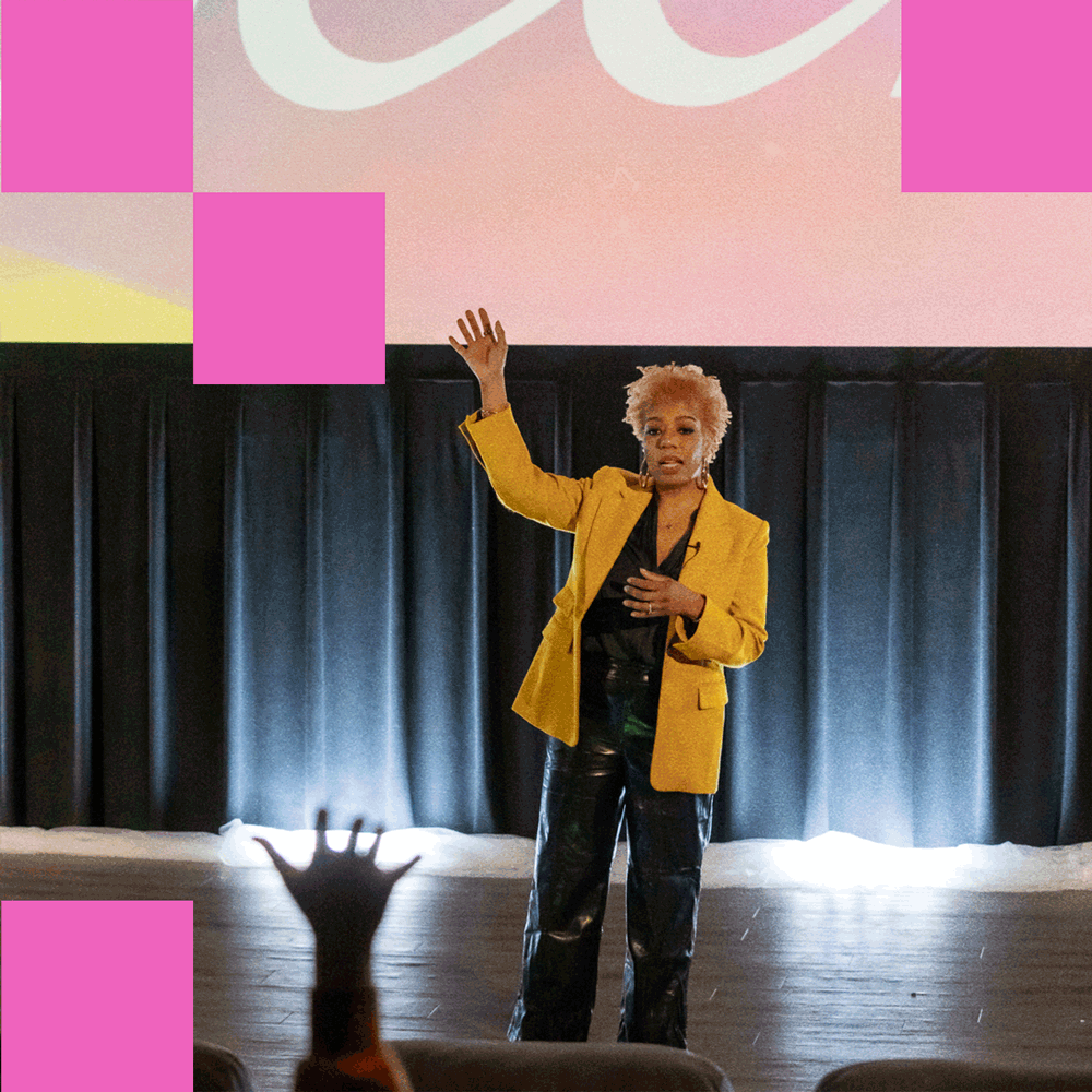 Black woman with short blond hair in a vibrant yellow blazer as an audience member raises their hand