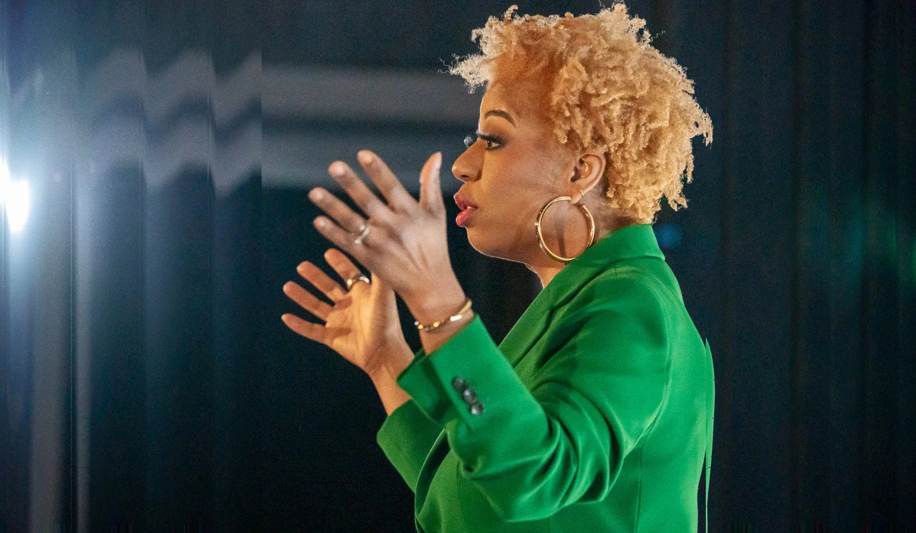 A woman with short, curly blonde hair is wearing a bright green blazer and large hoop earrings. She appears to be talking or gesturing with her hands while standing in front of a dark background.