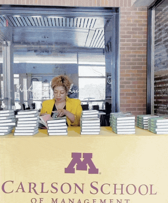 A woman in a yellow blazer organizing stacks of books at a table with a Carlson School of Management logo.
