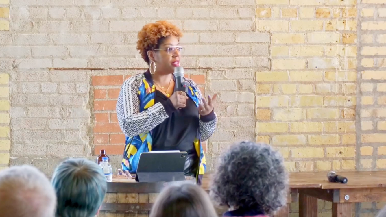 A woman with curly hair and glasses stands at a table, holding a microphone and speaking to an audience in front of a brick wall.