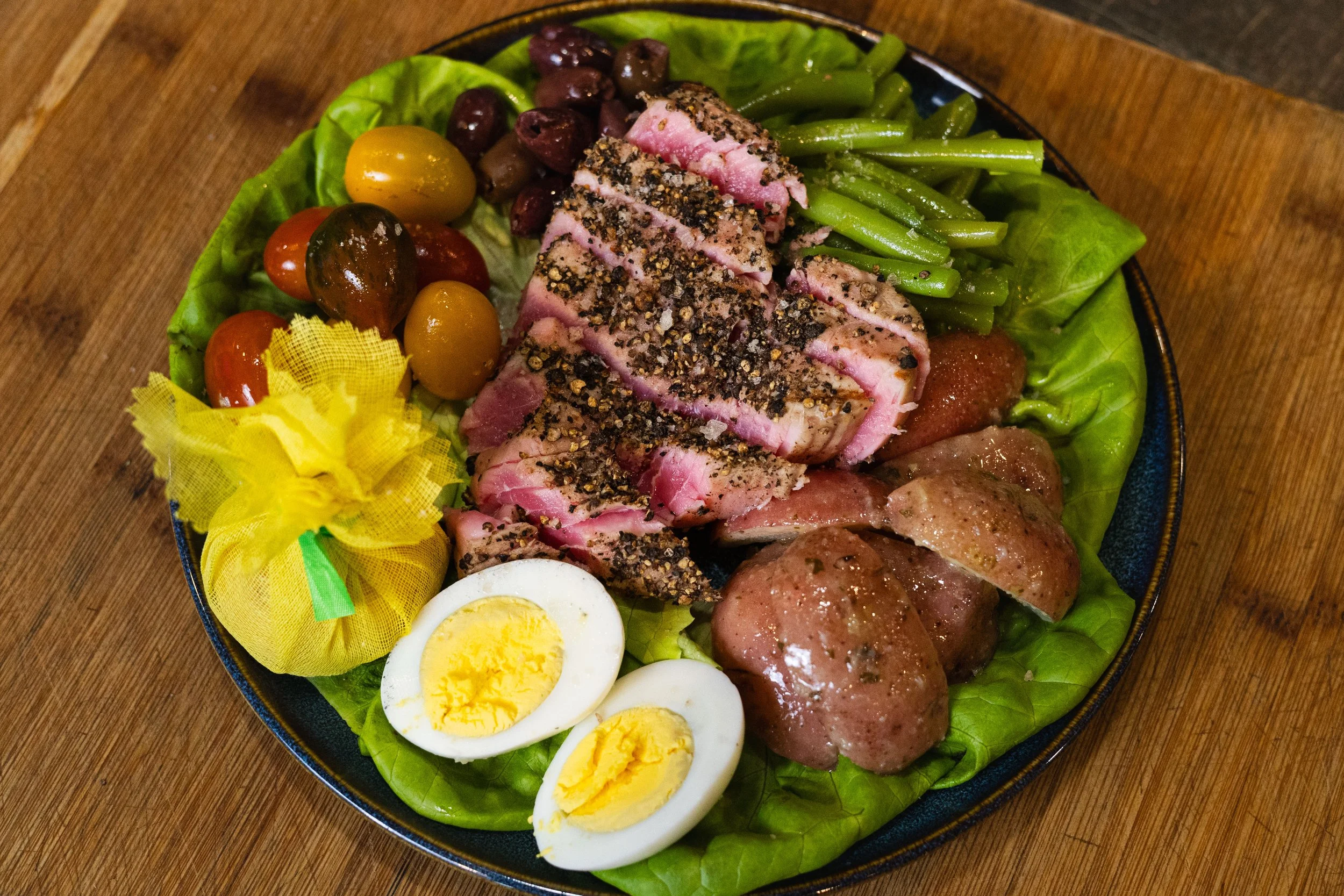 A plate of assorted foods including sliced seasoned tuna, boiled eggs, different colored cherry tomatoes, green beans, green leafy lettuce, stuffed grape leaves, and cooked sausages on a wooden surface.