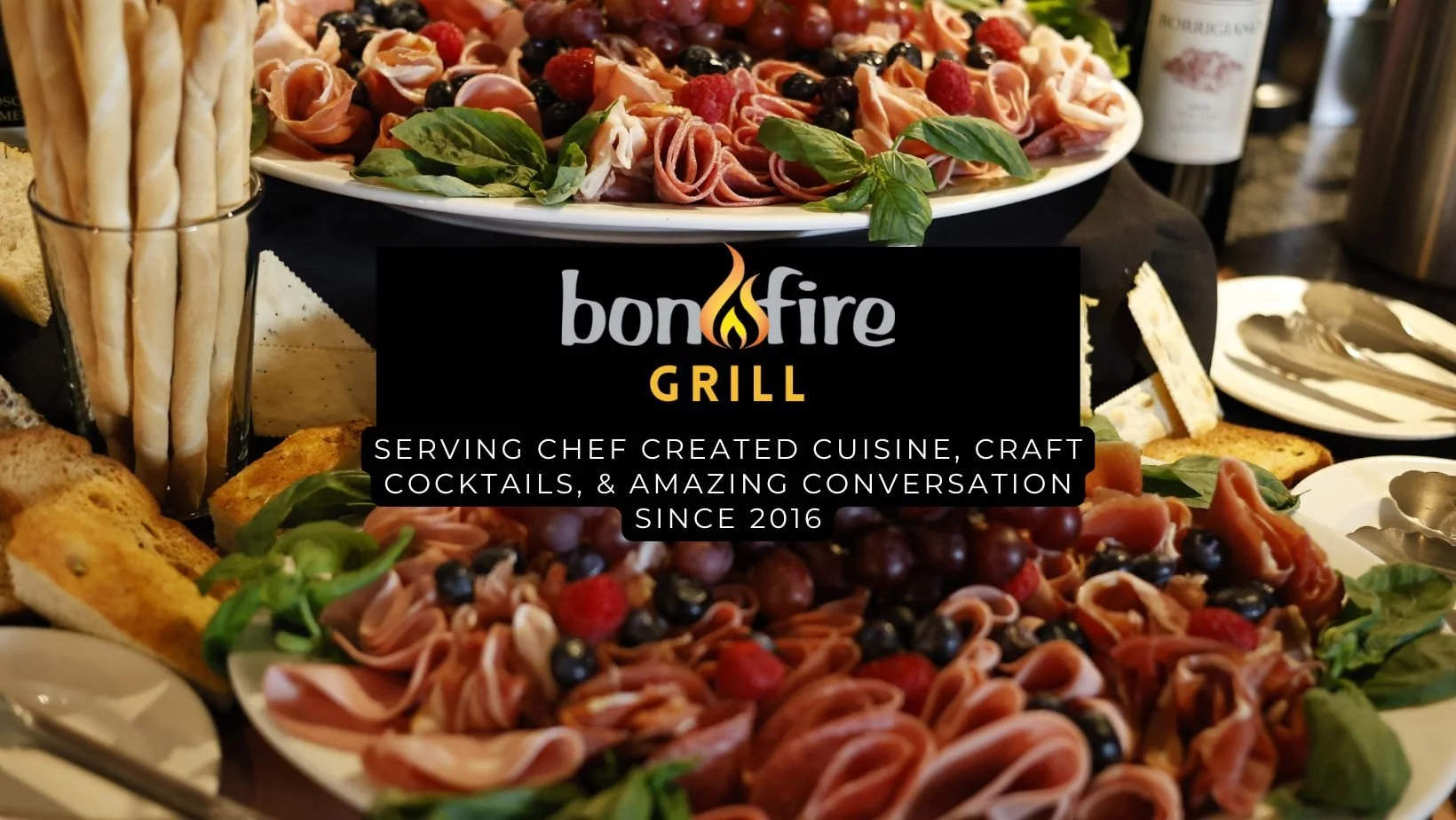 Plate of assorted cured meats, grapes, and fresh herbs, with slices of bread and breadsticks on the side, at Bonfire Grill.