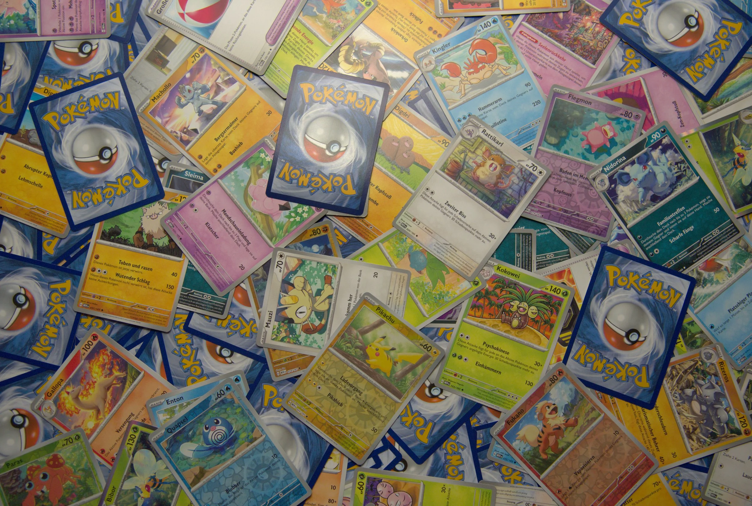 A pile of Pokémon trading cards scattered on a surface, including various Pokémon cards with colorful illustrations and different types, some facing up and others facing down.
