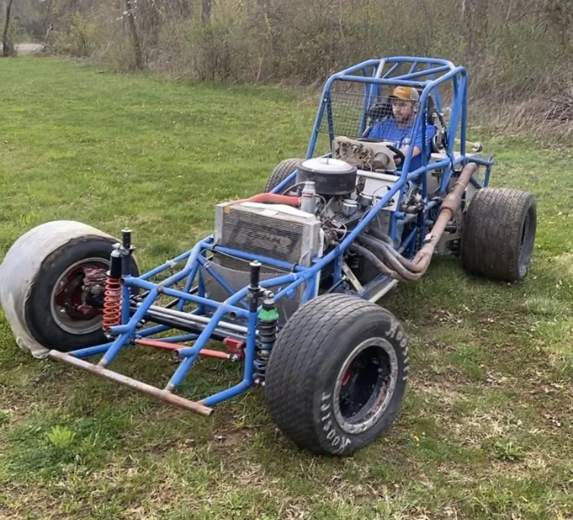 A person sitting in a blue off-road racing car on a grassy field.