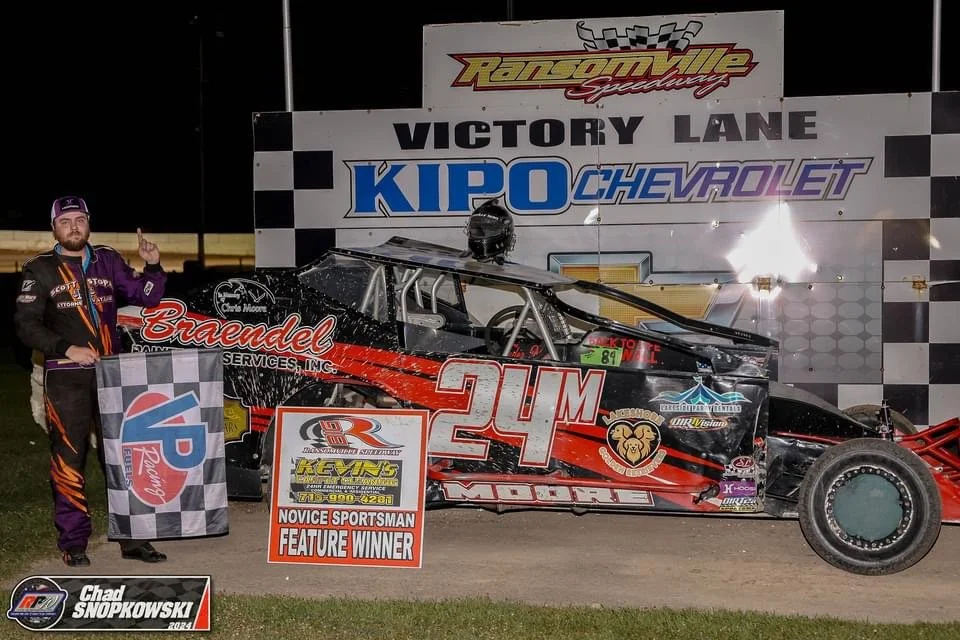 A race car driver standing next to his black and red race car at victory lane at a racetrack. The driver is holding a checkered flag and is pointing with his index finger. The race car has the number 24M on its side and various sponsor logos. A large