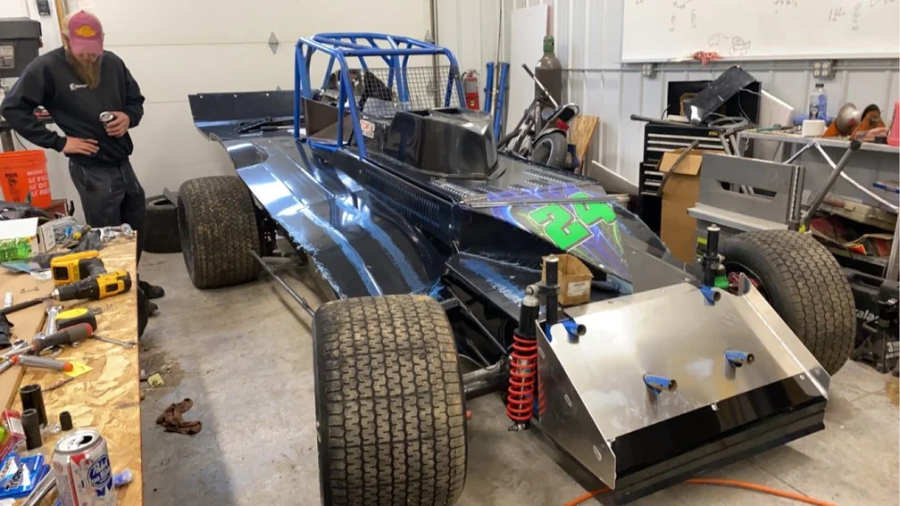 A racing car in a workshop with a person standing nearby. The car has large tires, a black body with blue and green accents, and a front wing. The person is wearing a black hoodie and pink cap. The workshop has tools and parts on a workbench and shel