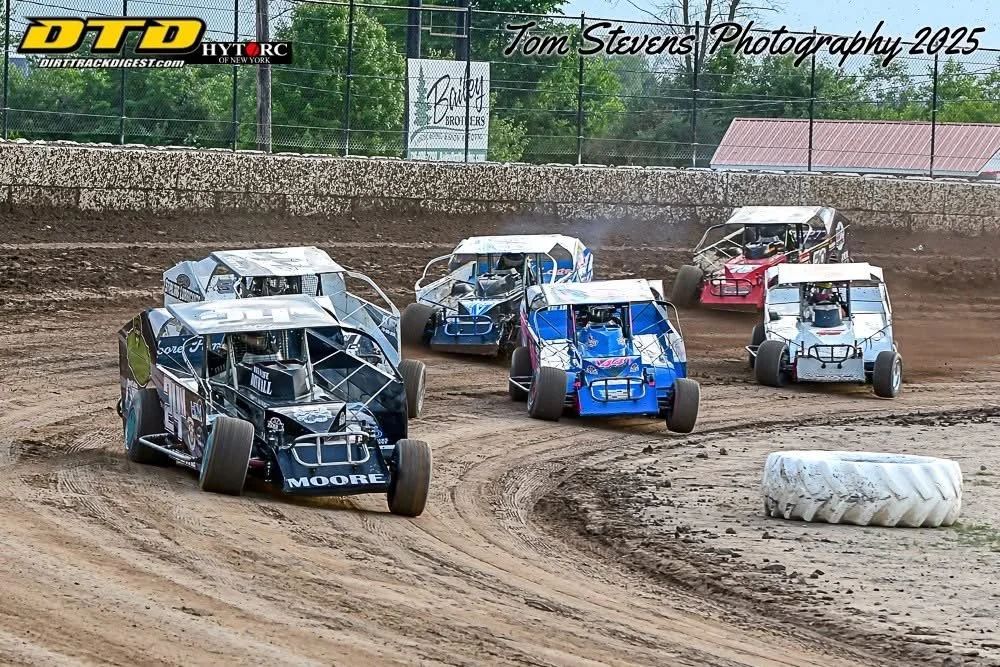 Five race cars on a dirt race track competing in a race, with a tire on the track and a fence with trees in the background.