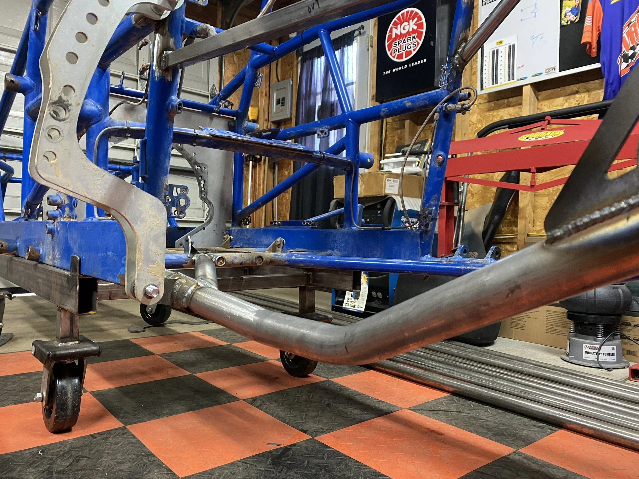 A racing go-kart frame with a metal pipe chassis on a checkered orange and black floor in a garage. There are tools and parts around, including a red cart, boxes, and posters on the wall.