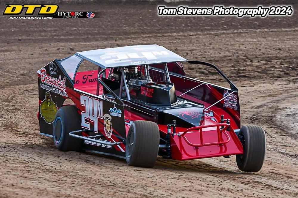 A red and black dirt track race car with the number 24M on the side, racing on a dirt track during daytime.
