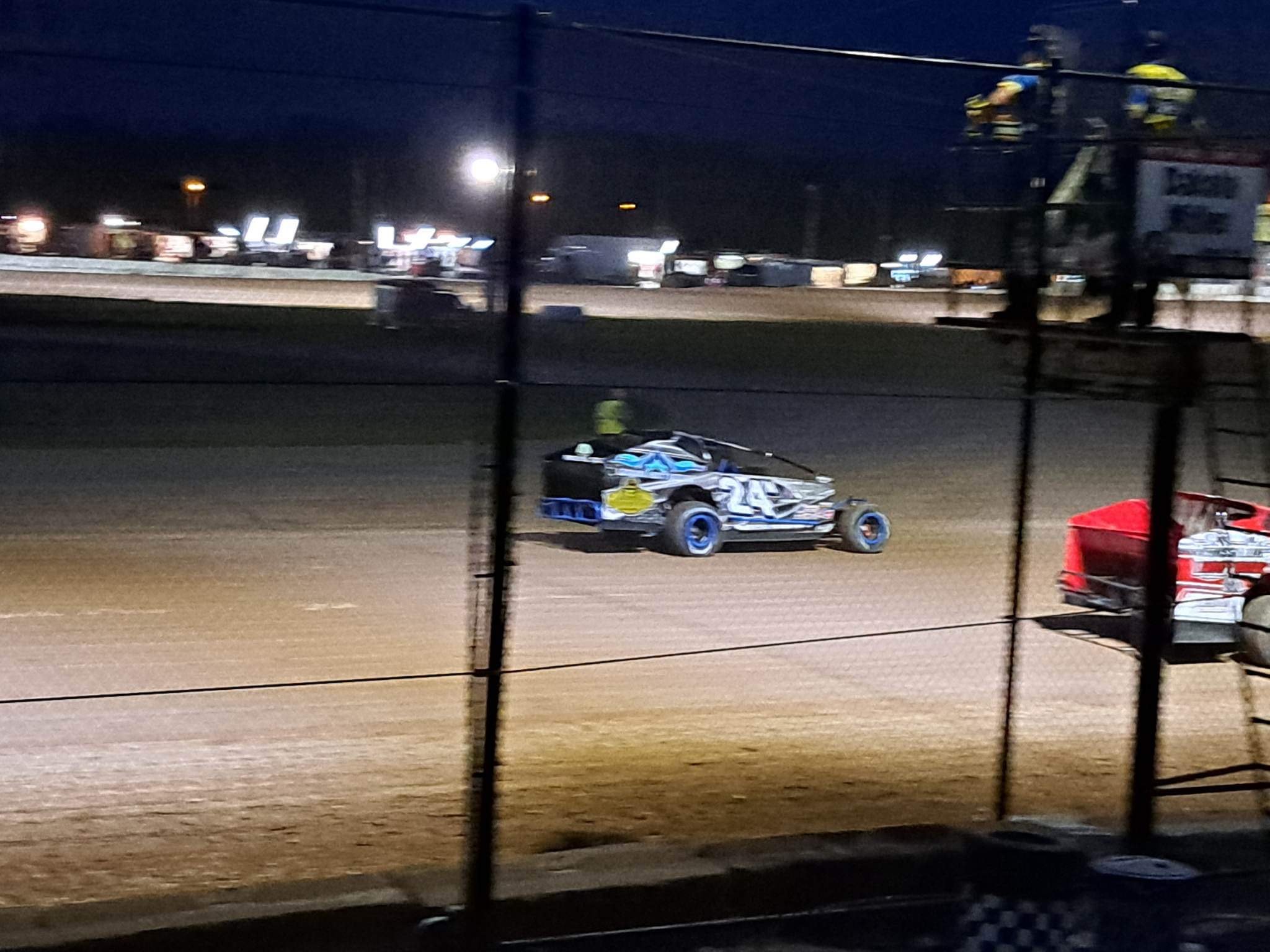 Nighttime dirt track racing scene with two race cars, one black with blue accents and the number 24, on the dirt track. Fencing in the foreground and spectators or crew members on a platform to the right.
