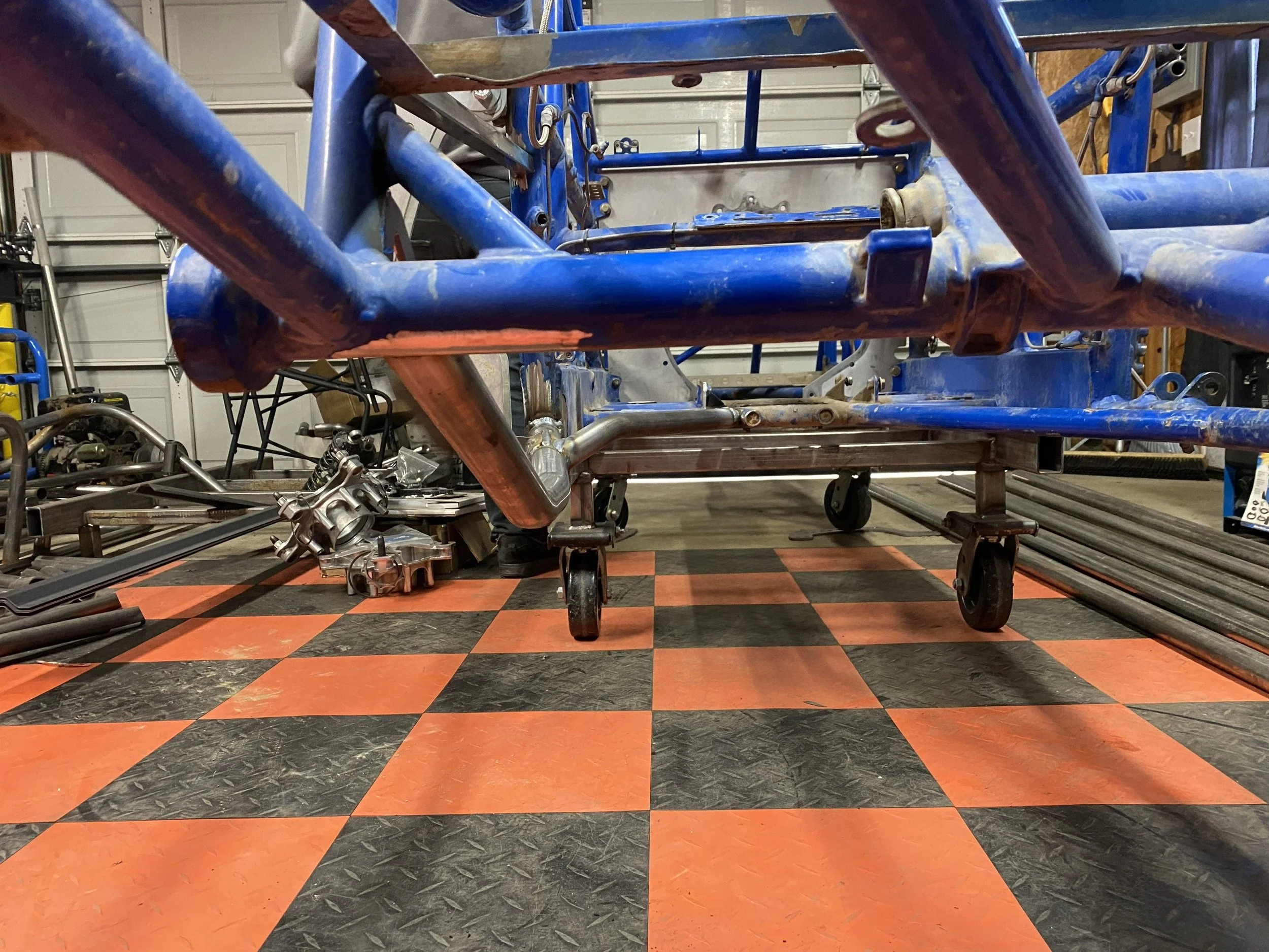 View of a garage or workshop with a racing car chassis on a wheeled cart, showing blue metal frames and parts, with tools and engine components nearby. The floor has a black and orange checkered pattern.