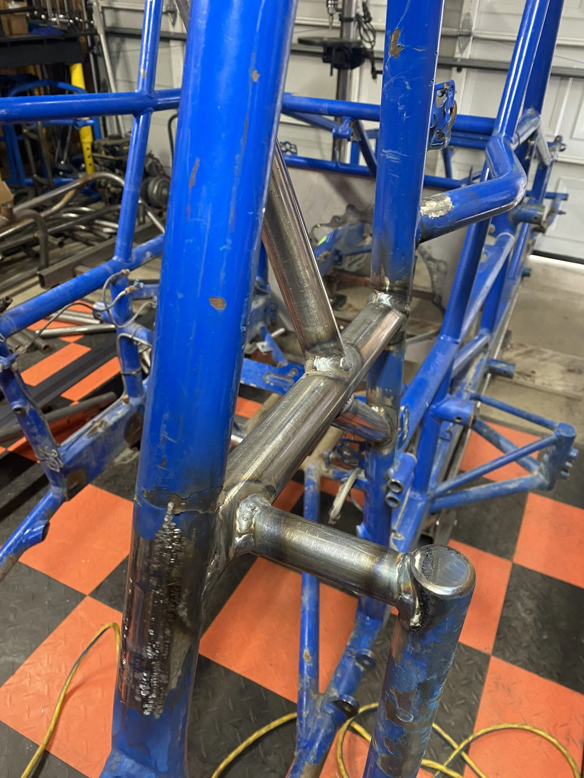 Metal frame of a bicycle or similar structure under construction, with blue paint and welds visible, in a workshop or garage.