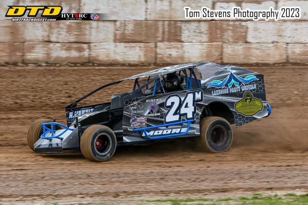 A black and blue dirt track race car with the number 24M, kicking up dirt as it speeds on a dirt racetrack with a brick wall in the background. The car has advertisements and sponsorship decals, including Lakeside Party Rentals, and is equipped with 
