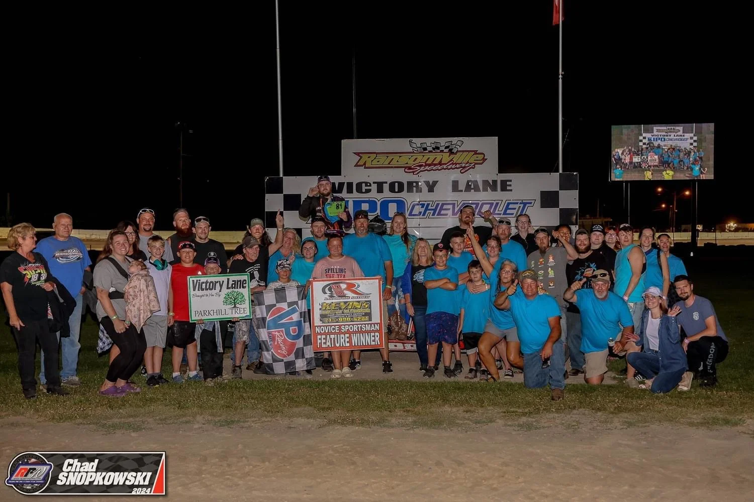 A large group of people celebrating at a racing victory event at Victory Lane Speedway, with a night sky and race track in the background. The group includes men, women, and children, some wearing blue shirts, holding signs, and raising their arms in
