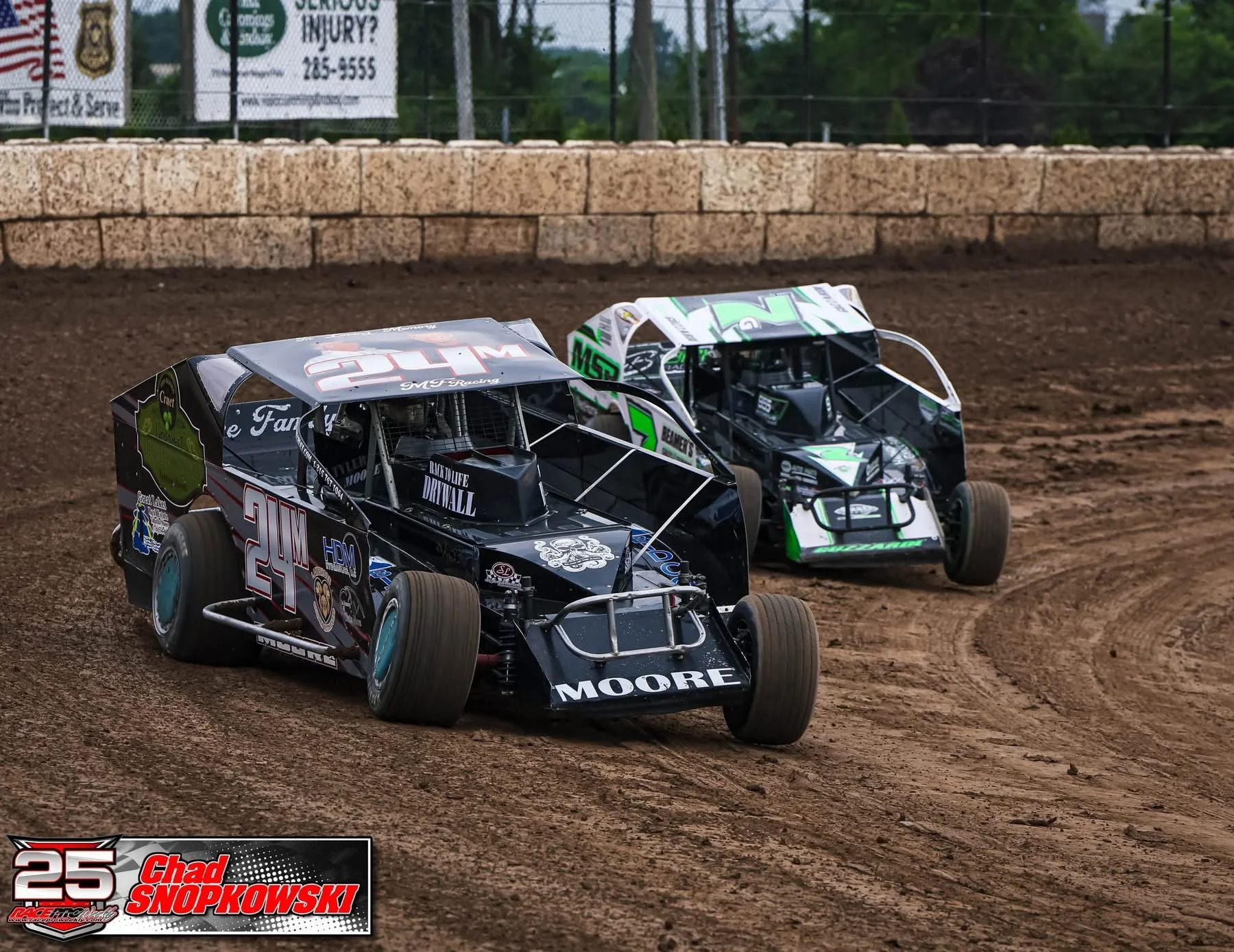 Two race cars on a dirt track, number 27 in front, number 55 behind, with a brick wall and green trees in the background during a dirt track race.