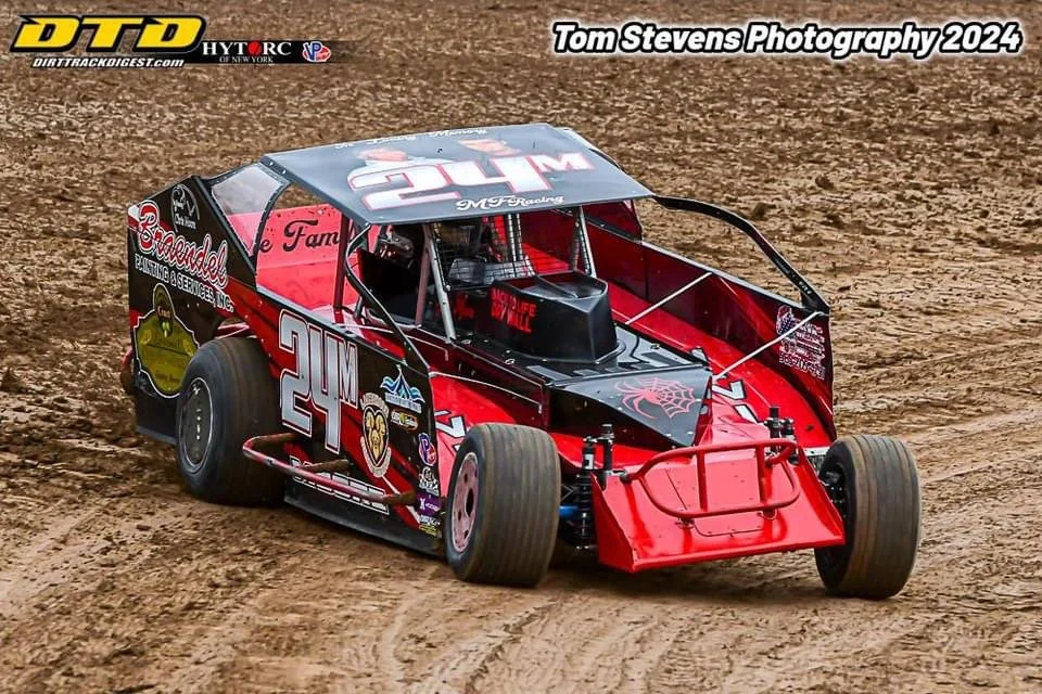Red and black dirt race car with the number 24M on the side, racing on a dirt track. The car has several sponsor logos and a wing on top, with muddy tires and a dirt surface.