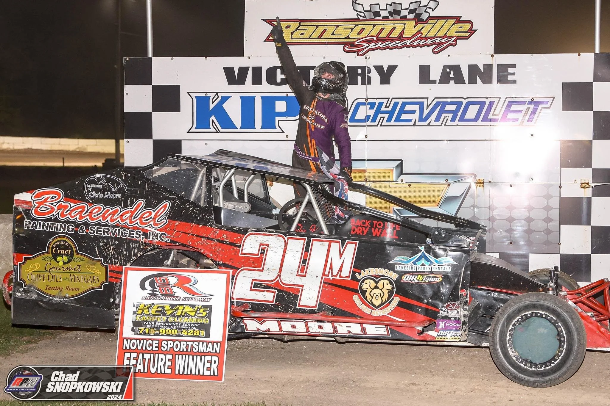 Race car driver celebrating on the victory lane of Ransomville Speedway after winning the novice sportsman feature. The driver, wearing racing gear and helmet, stands with one arm raised. The car, number 24M, is black and red with various sponsor log