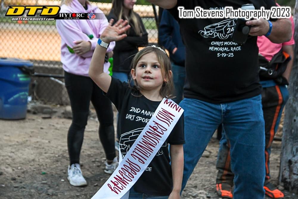 A young girl raising her right hand at a dirt track event, wearing a sash that reads 'Mini,' with a black T-shirt promoting the 2nd Annual Dirt Sport race. Behind her, several people including a woman in black leggings and a pink jacket, and a man ho