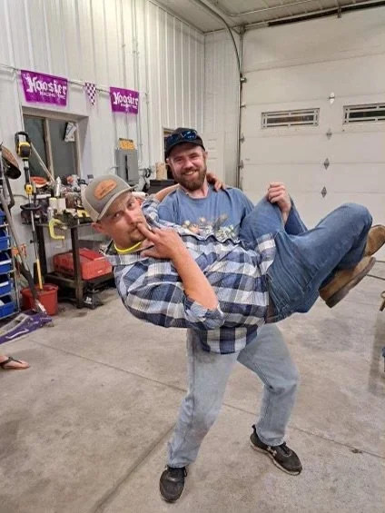 Two men in a garage, one is holding the other who is laying horizontally, both smiling.