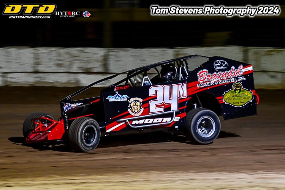 A black and red sprint car with the number 24M racing on a dirt track, with advertisements and sponsor logos on the car, during a night race.