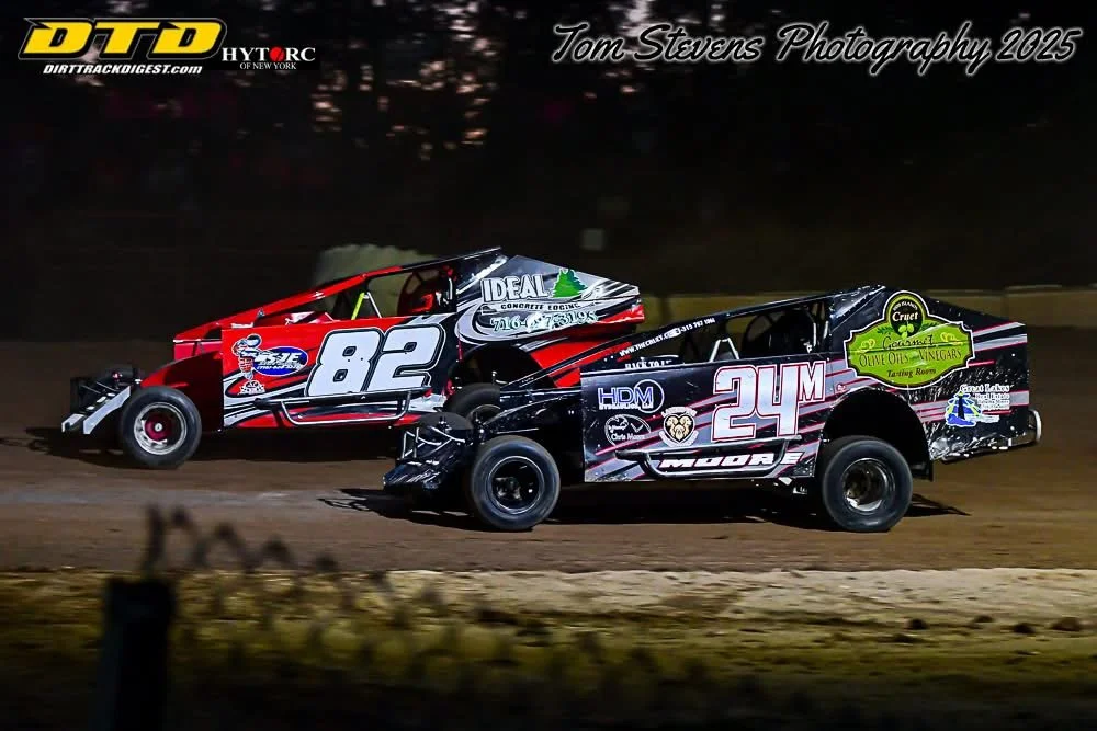 Two dirt track racing cars, number 82 red and black and number 24 black and gray, racing on a dirt track at night with a dark background and some blurred foreground.