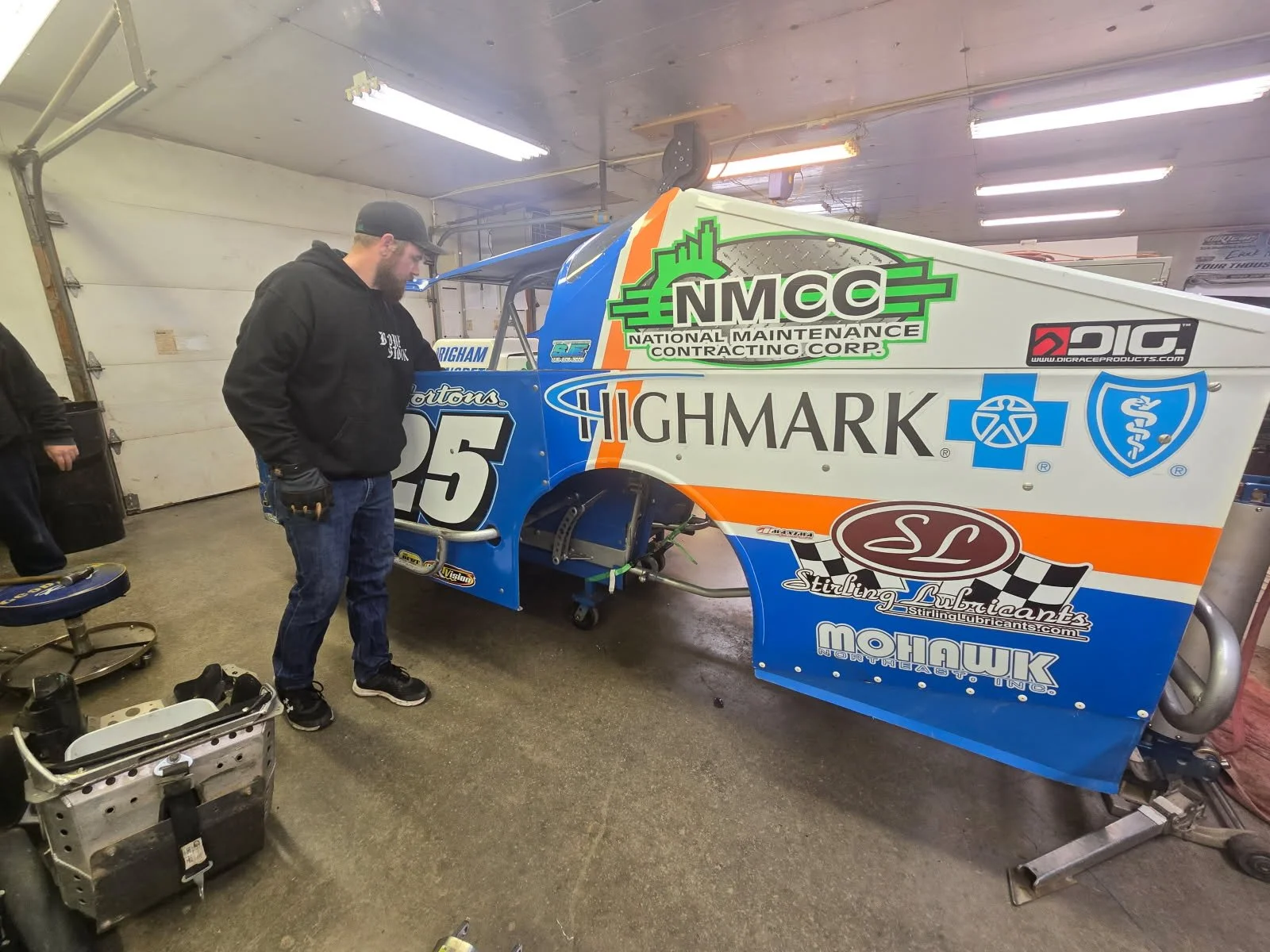 A man inspecting a race car in a garage. The car's body is decorated with various sponsor logos and a number 25.