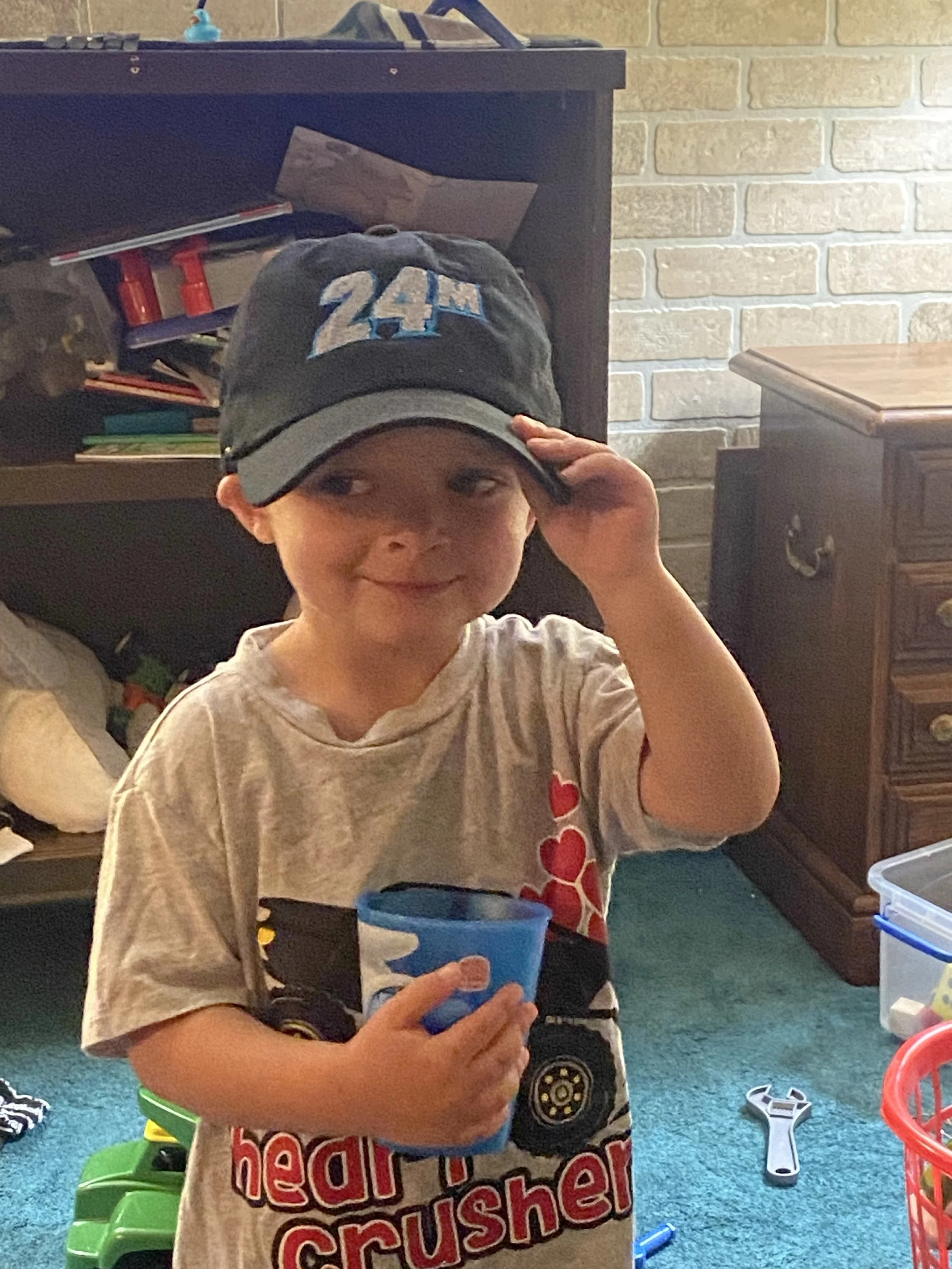 A young boy wearing a black baseball cap with the number 24 in blue, holding a blue cup, and smiling while looking to the side in a room with toys and furniture.