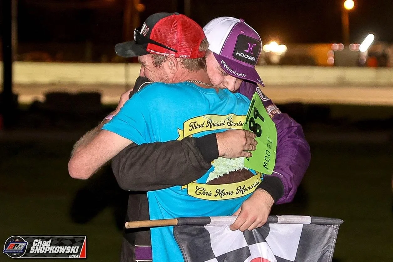 Two race car drivers embrace on a racetrack at night, one wearing a blue shirt with a yellow ribbon and the other wearing a purple racing suit and cap, holding a flag, with bright lights in the background.