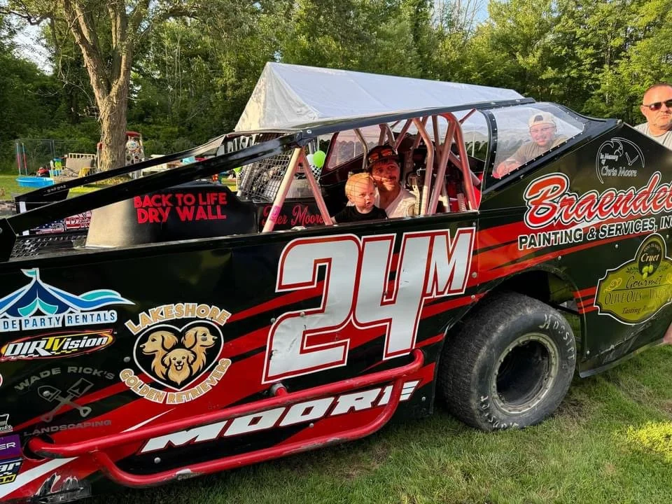 A black and red race car with the number 24 and several sponsor logos, parked on grass with trees in the background. Two people are inside the car, and a man and a child are sitting behind it. A man with sunglasses is standing nearby.
