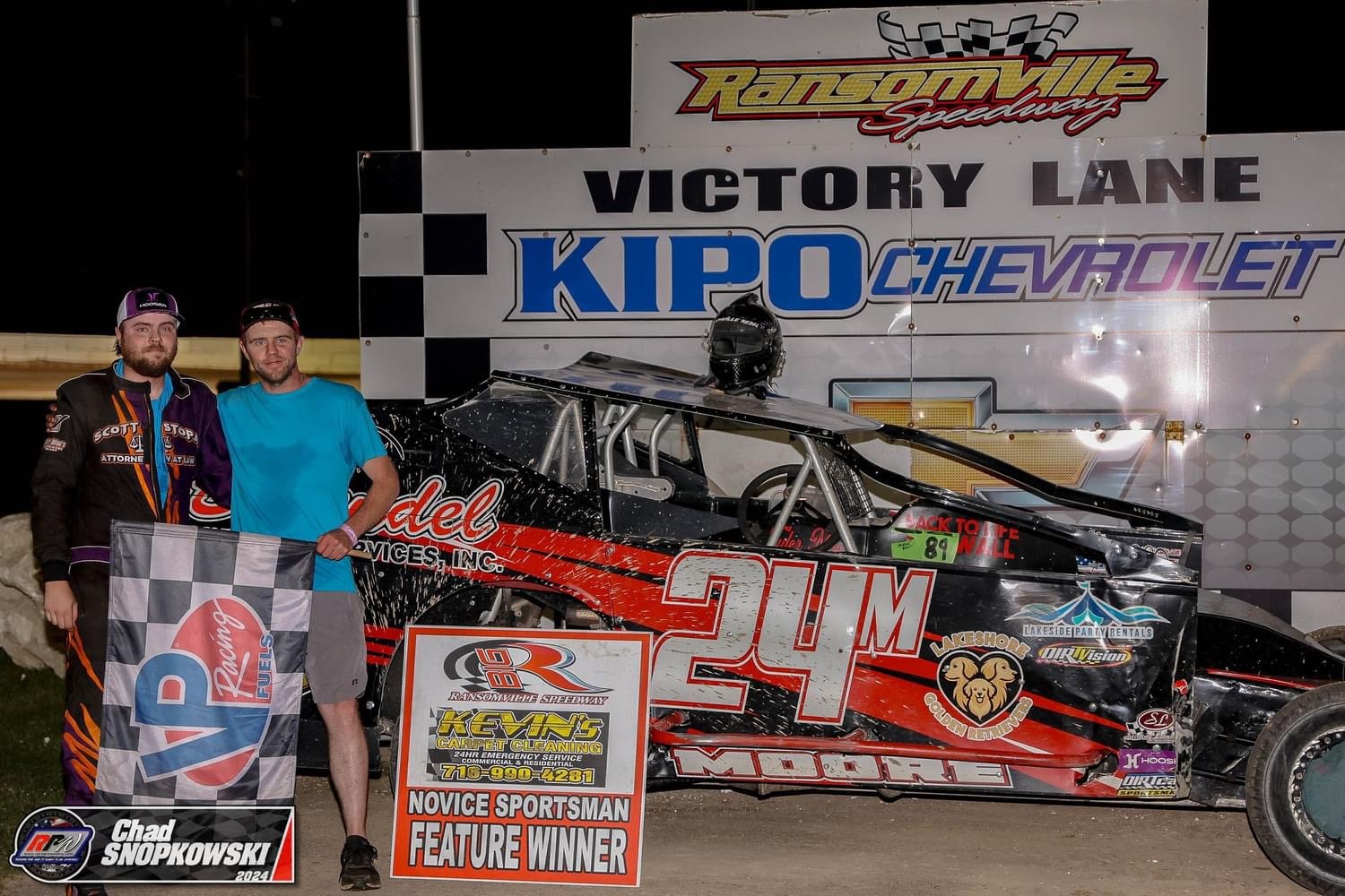 Two men standing next to a racing car at Ransomville Speedway victory lane, with a large sign behind them. One man is holding a checkered flag, and one has his arm around the other. The car has the number 21M and various sponsor logos. A helmet is pl